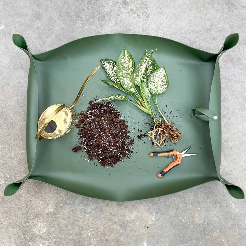 Un grand tapis de rempotage vert sombre en simili cuir avec une plante, du terreau, un arrosoir et un sécateur